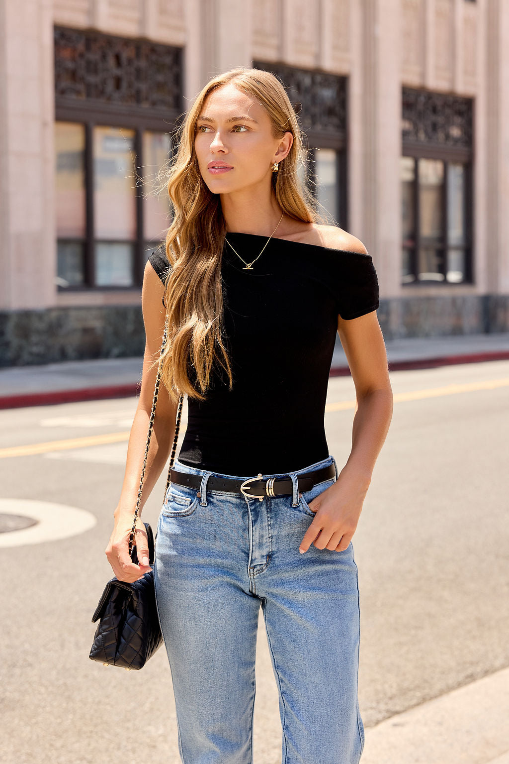 Woman with long hair wears a Black Asymmetrical Ribbed Top and jeans, holding a black bag on a city street.