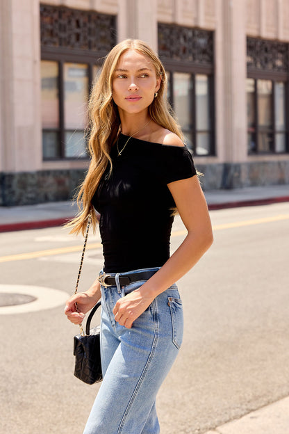 Woman with long blonde hair in a Black Asymmetrical Ribbed Top and blue jeans, walking on a city street holding a small black purse.