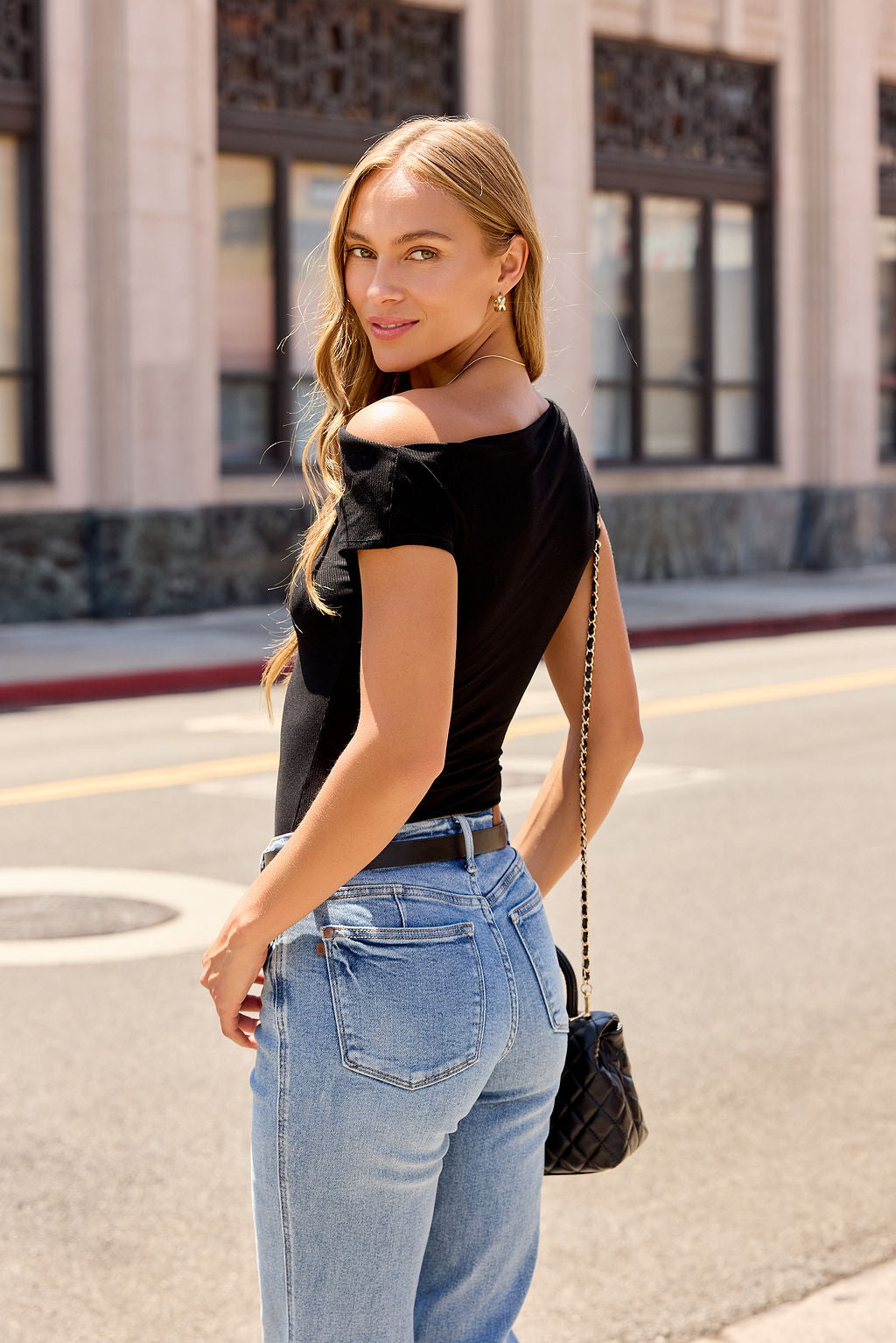 Woman with long blonde hair wears a Black Asymmetrical Ribbed Top and jeans, holding a black purse on a city street.