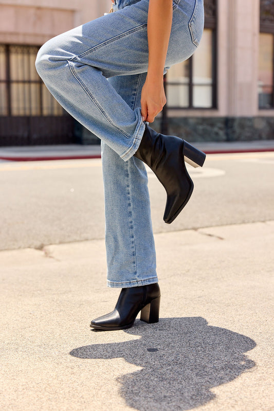 Person in blue jeans adjusts MIA Sidonie Leather Booties - Black | Pre Order on a city sidewalk, shadow visible on the ground.
