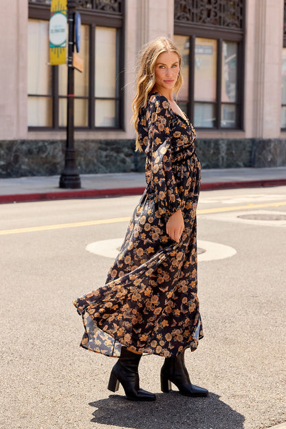 Woman wears the Maple Floral Maxi Dress - Black | Pre Order with black boots, posing elegantly on a city street framed by buildings.