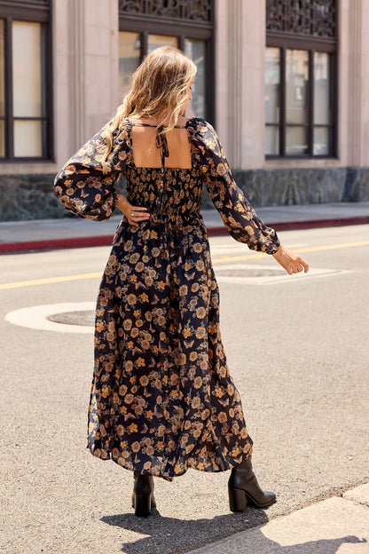 A woman in the Maple Floral Maxi Dress - Black | Pre Order and black boots walks across a city street, facing away from the camera.