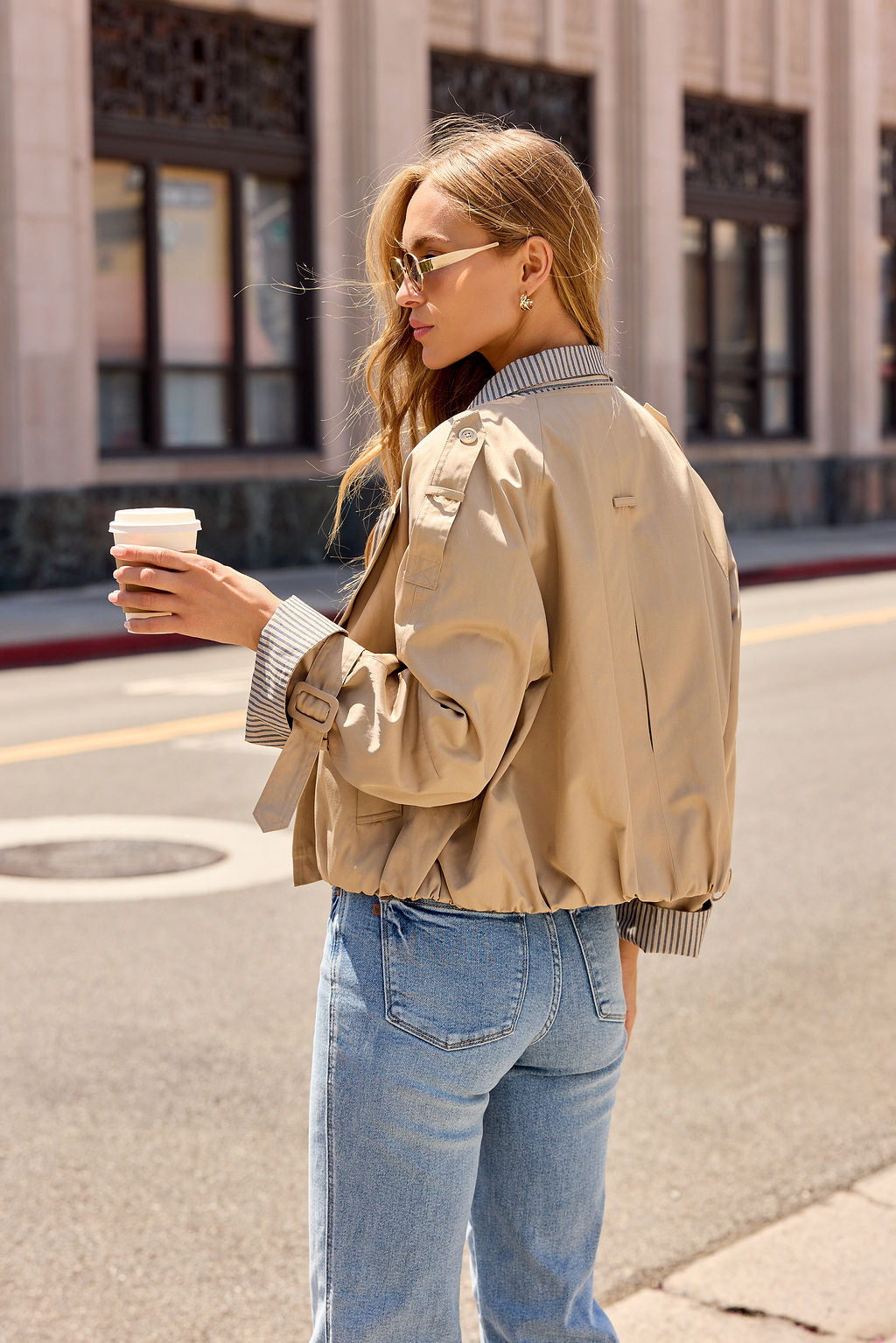 Woman in sunglasses and jeans wearing the Contrast Striped Shirt Jacket - Camel, holding coffee on a city street.