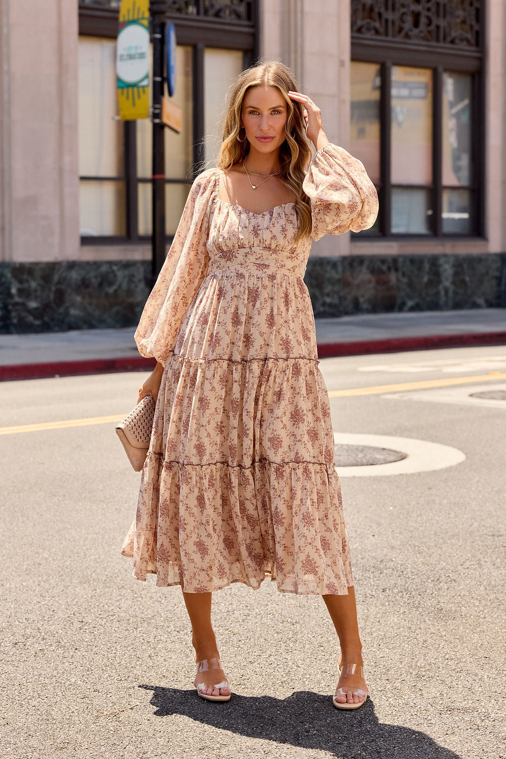 Woman in the Sweetheart Floral Print Midi Dress - Sand stands on a sunny city street, holding a beige clutch.
