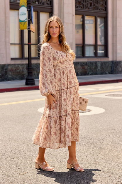 Woman wearing the Sweetheart Floral Print Midi Dress - Sand, sheer sleeves, tiered hem, clear-heeled sandals, and a beige clutch.
