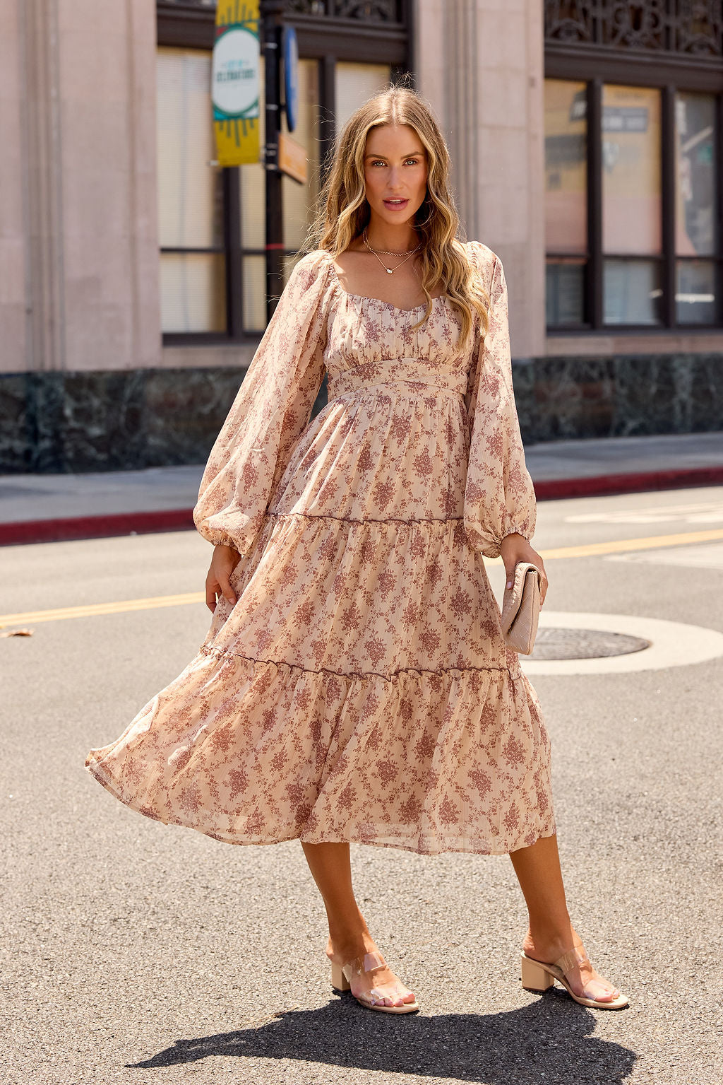 Woman in the Sweetheart Floral Print Midi Dress - Sand and sandals stands on a city street holding a clutch, buildings behind her.