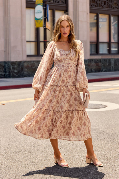Woman in the Sweetheart Floral Print Midi Dress - Sand and sandals stands on a city street holding a clutch, buildings behind her.