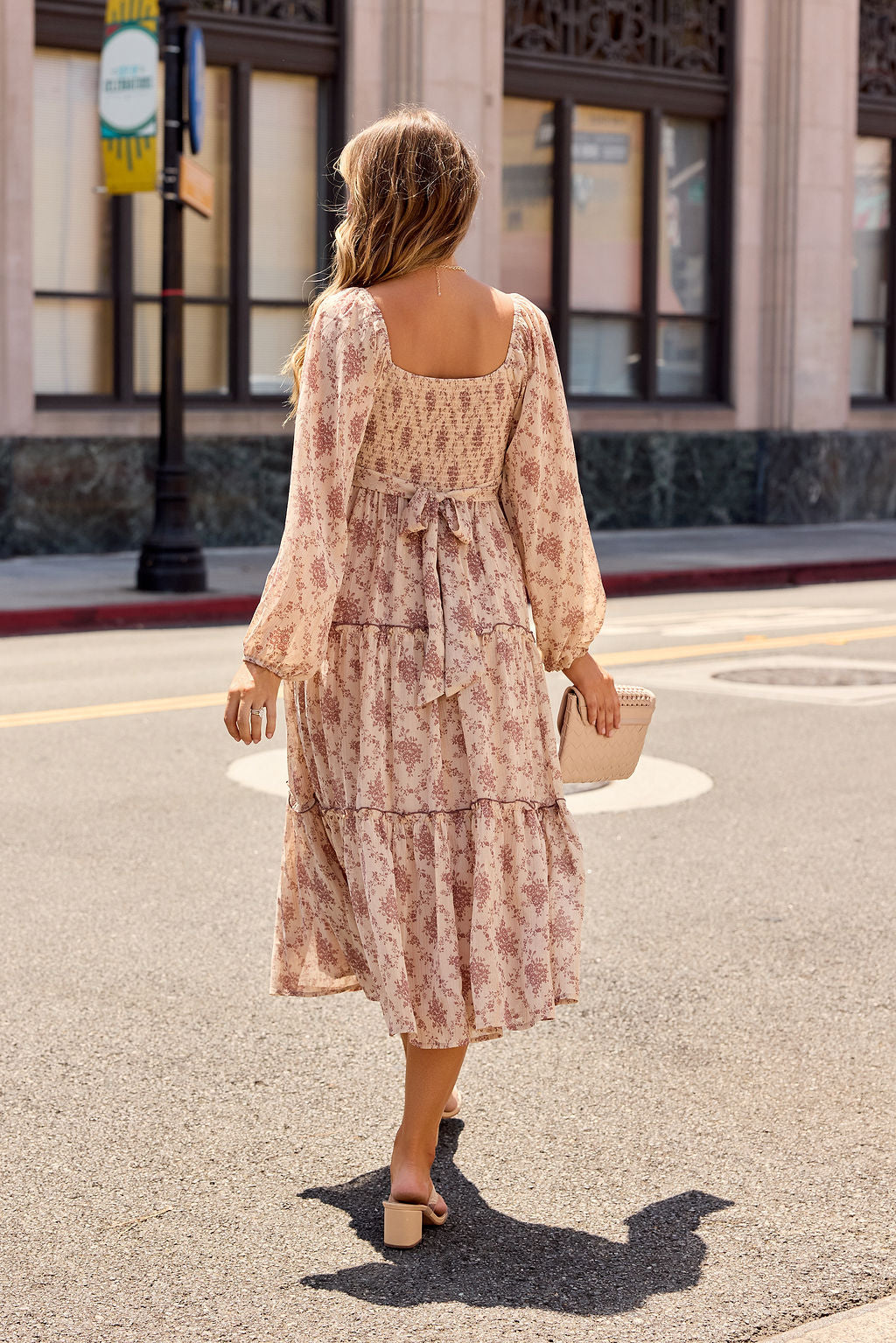A woman in the Sweetheart Floral Print Midi Dress - Sand and heels walks a city street, clutch in hand, back to the camera.