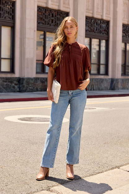 Woman on city street wearing Puff Sleeve Tie Front Top in brown, blue jeans, beige heels, and holding a white clutch.