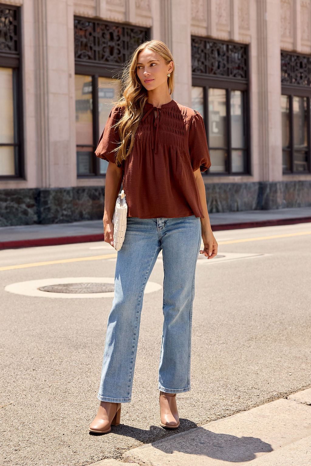Woman in a brown Puff Sleeve Tie Front Top, light blue jeans, and tan heels stands on a city street holding a white clutch.