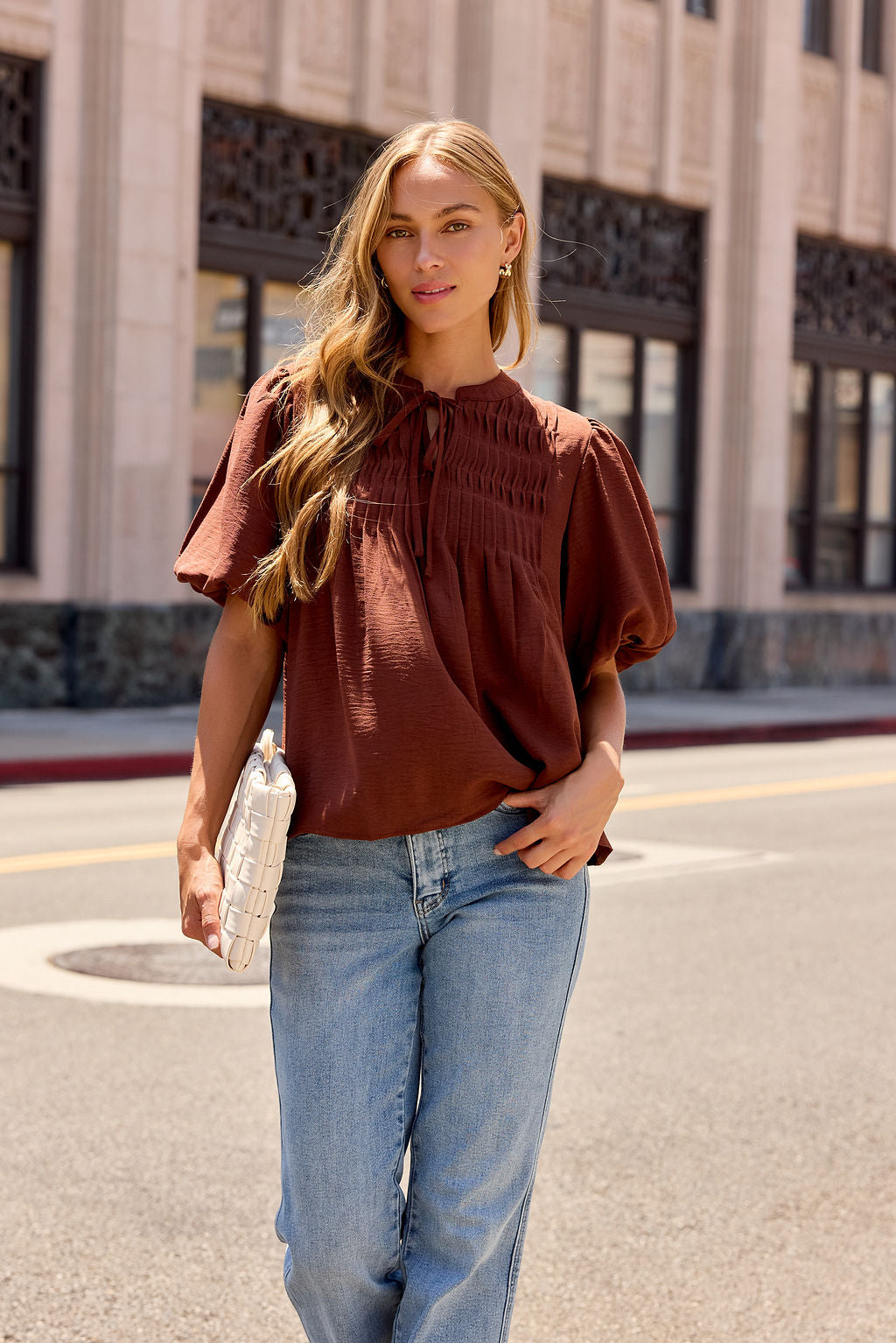 Woman stands on a city street in a Puff Sleeve Tie Front Top - Brown and jeans, holding a white clutch purse, building behind her.