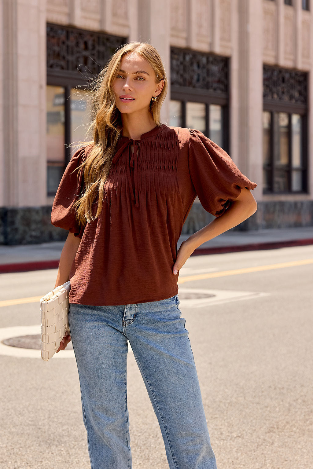 Woman in a brown Puff Sleeve Tie Front Top and blue jeans stands on a city street, holding a white woven clutch.