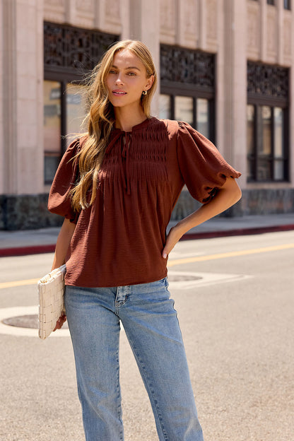 Woman in a brown Puff Sleeve Tie Front Top and blue jeans stands on a city street, holding a white woven clutch.