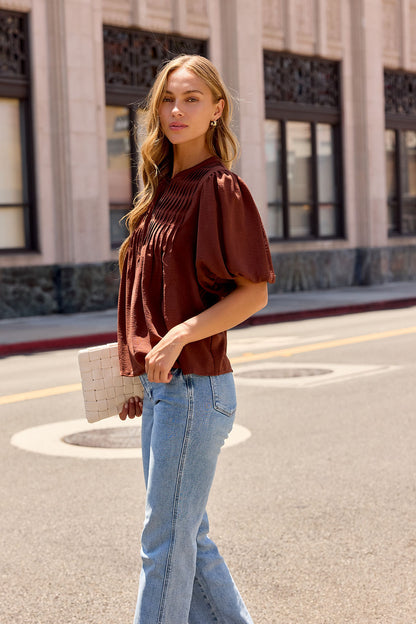 A woman in a brown Puff Sleeve Tie Front Top and blue jeans stands on a city street, holding a white clutch, looking at the camera.