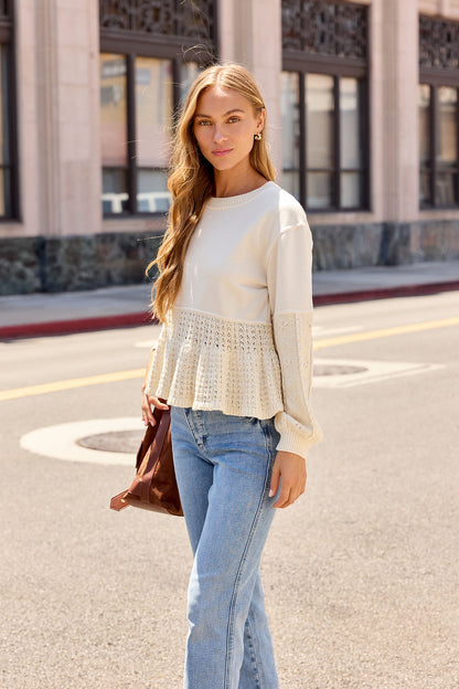 Woman with long hair in a Wren Textured Sweater - Cream stands on a city street, holding a brown bag and looking at the camera.