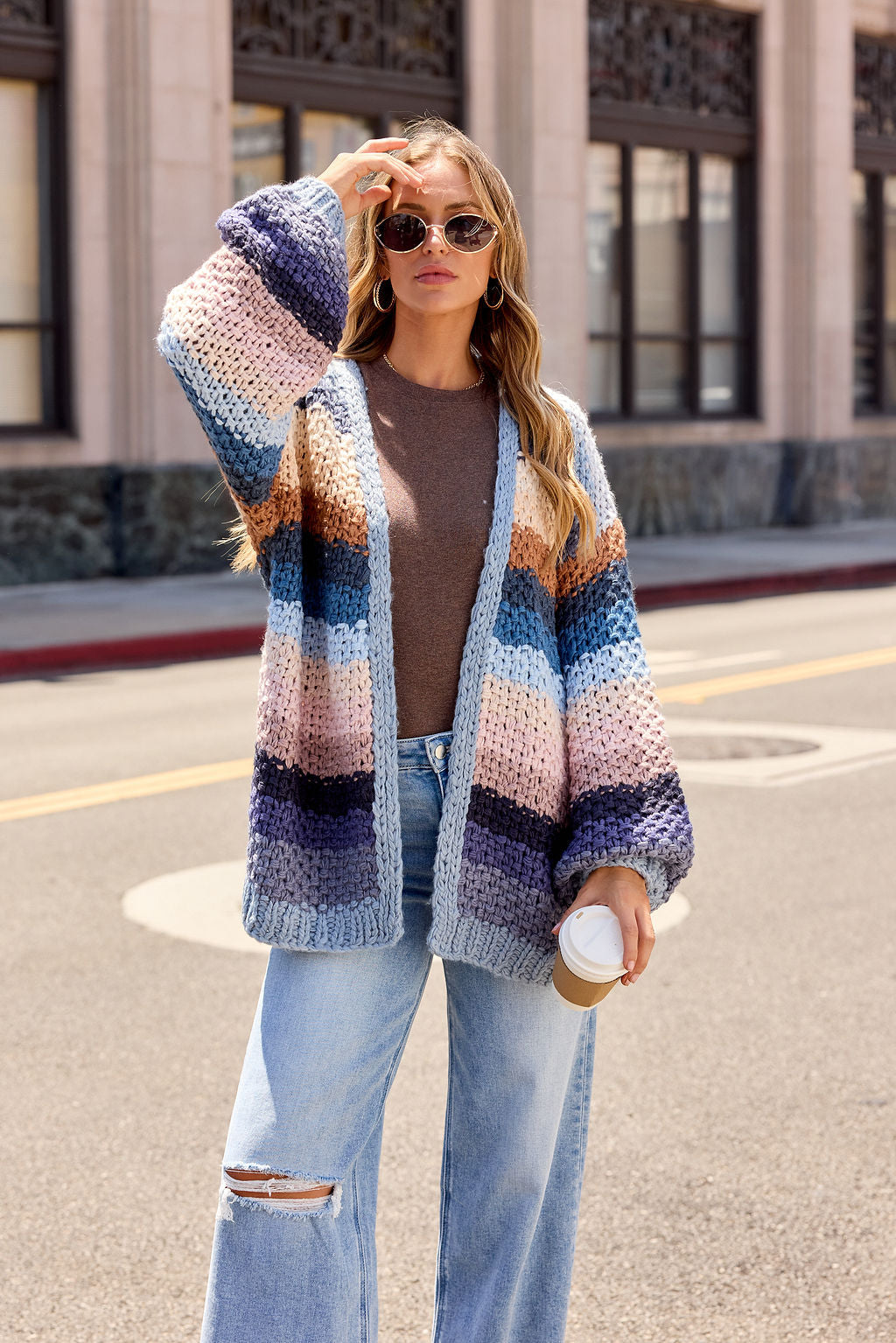 Woman in sunglasses wears the Multi Blue Stripe Crochet Cardigan, brown top, and jeans, holding coffee on a city street.