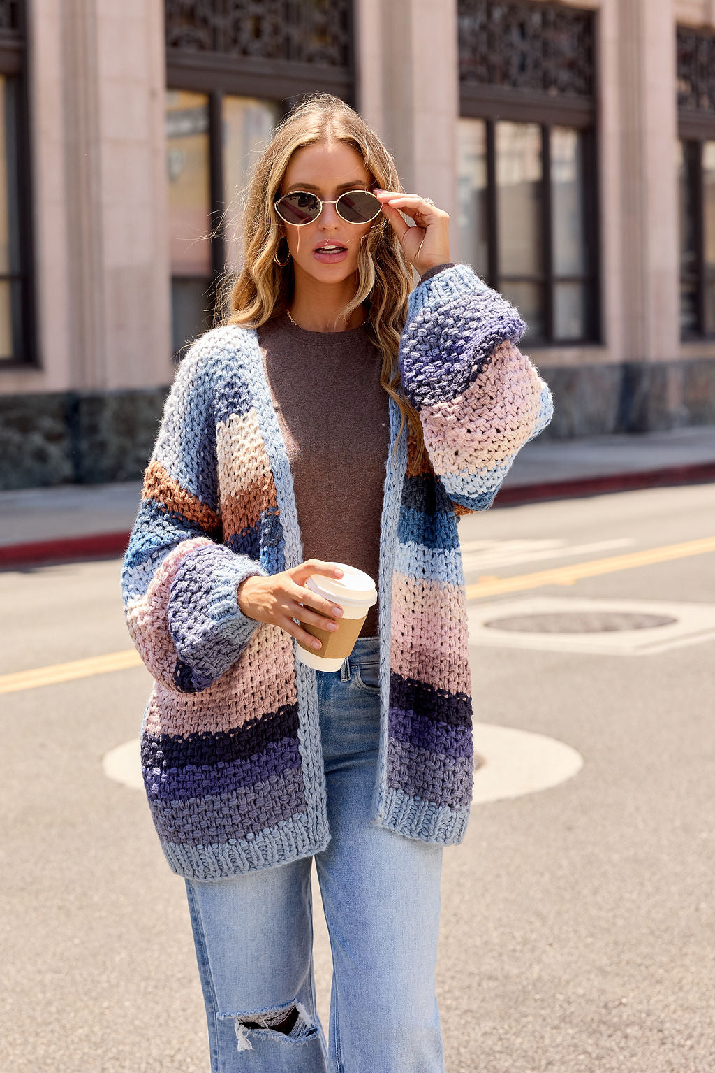 A woman in a Multi Blue Stripe Crochet Cardigan - Pre Order and sunglasses holds coffee on a city street.