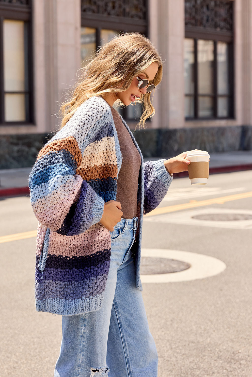 Woman in a Multi Blue Stripe Crochet Cardigan, sunglasses, and jeans walks outside holding coffee on a city street.