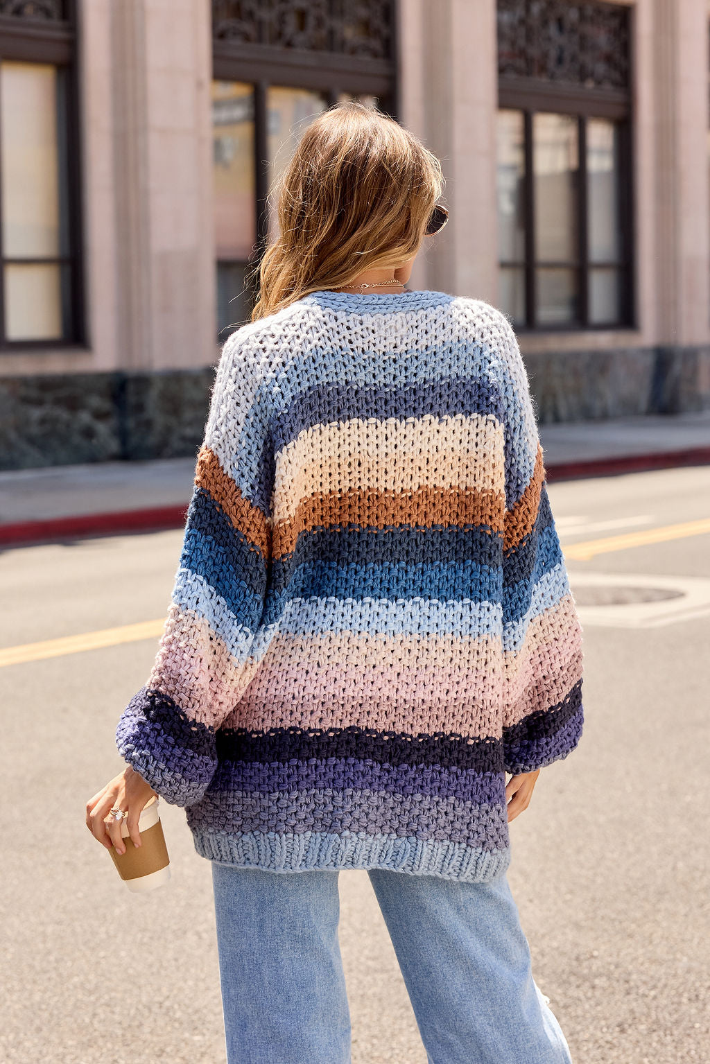 A person in a Multi Blue Stripe Crochet Cardigan holds a coffee cup on a city street, facing away from the camera.