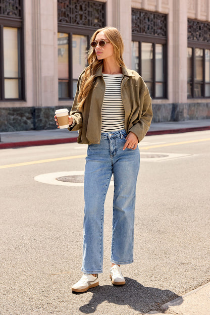 Woman in sunglasses, striped top, and jeans wears the Suede Bubble Hem Jacket - Olive while crossing a sunny city street with coffee.