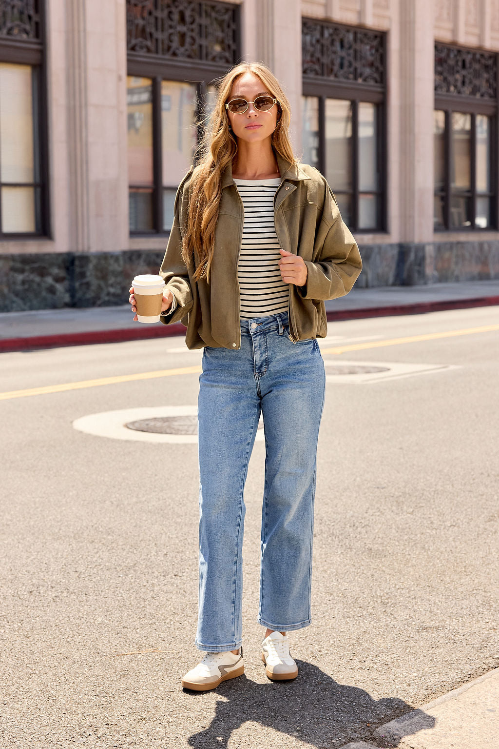 Woman in sunglasses, striped shirt, and jeans layers with the Suede Bubble Hem Jacket - Olive, holding coffee on a city street.