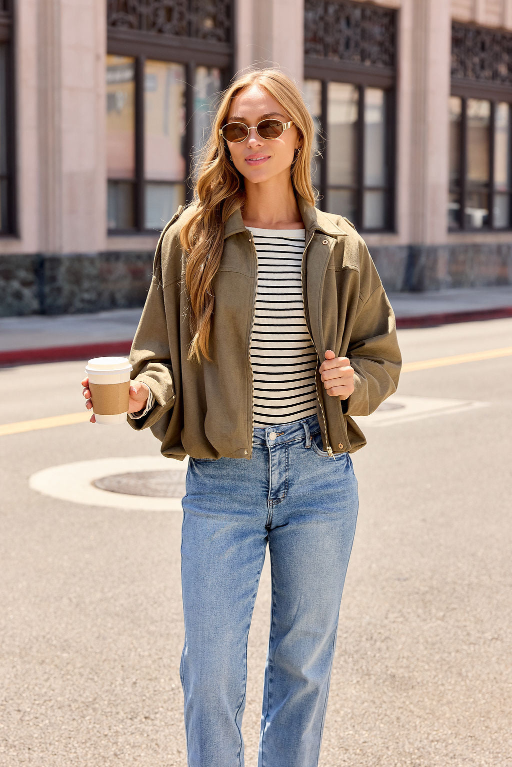 Woman in sunglasses, striped shirt, and Suede Bubble Hem Jacket - Olive holding coffee—perfect fall city layering.