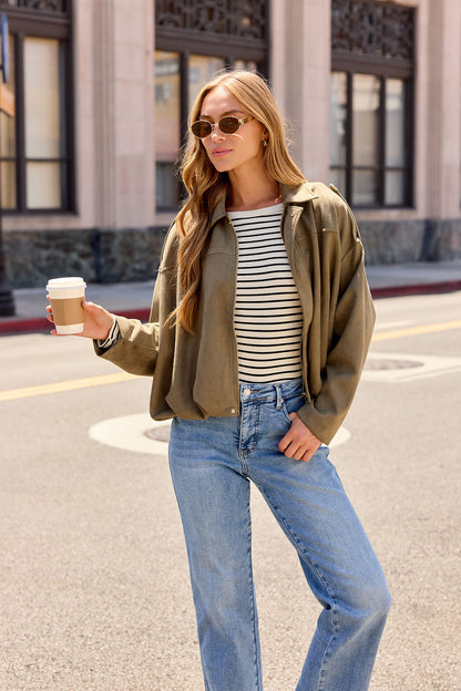 Woman in sunglasses, striped shirt, and jeans wears the Olive Suede Bubble Hem Jacket, holding coffee on a sunlit city street.