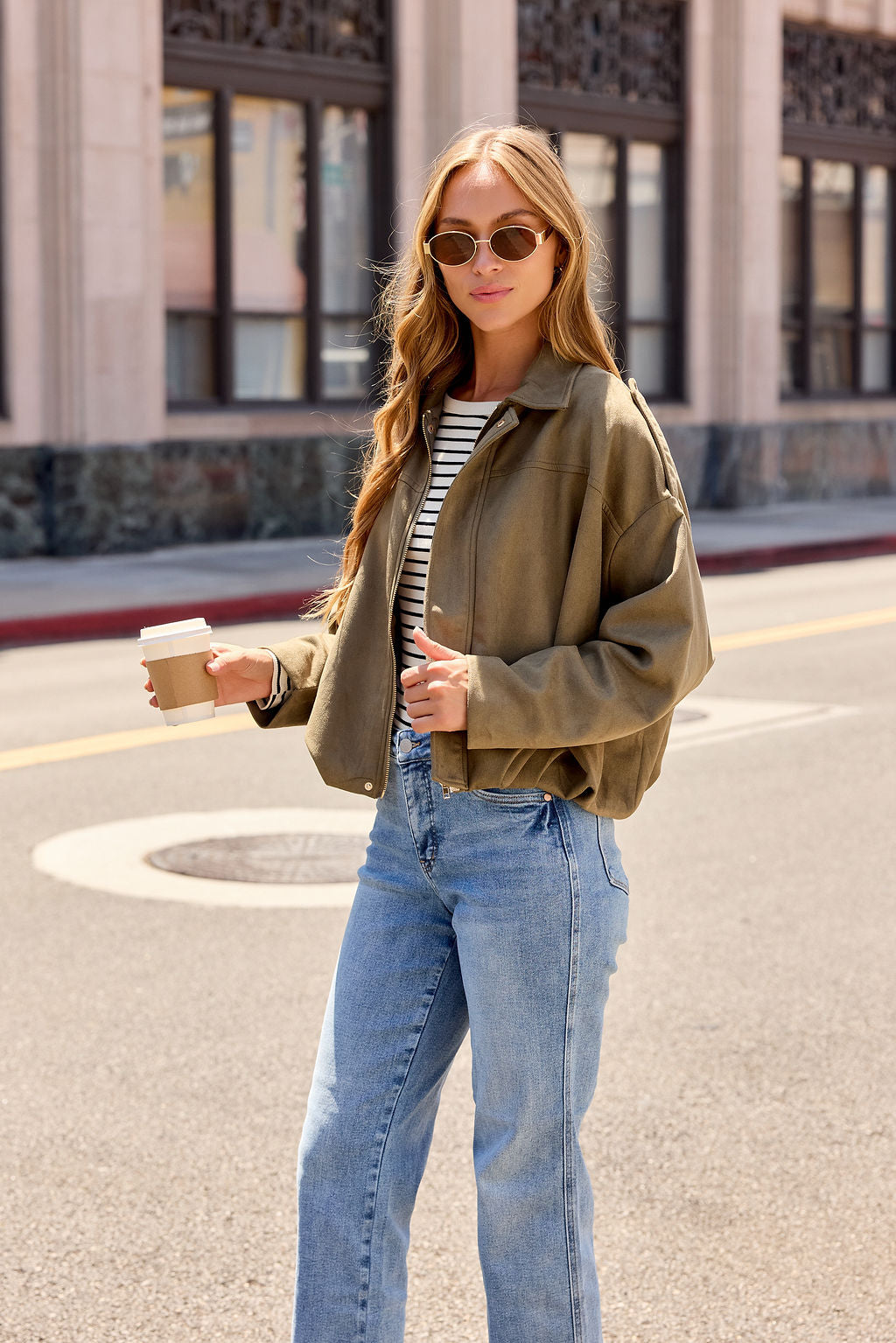 Woman in sunglasses, striped shirt, jeans, and the Suede Bubble Hem Jacket - Olive holds a coffee on a city street in daylight.