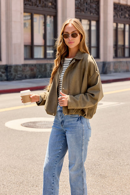 Woman in sunglasses, striped shirt, jeans, and the Suede Bubble Hem Jacket - Olive holds a coffee on a city street in daylight.