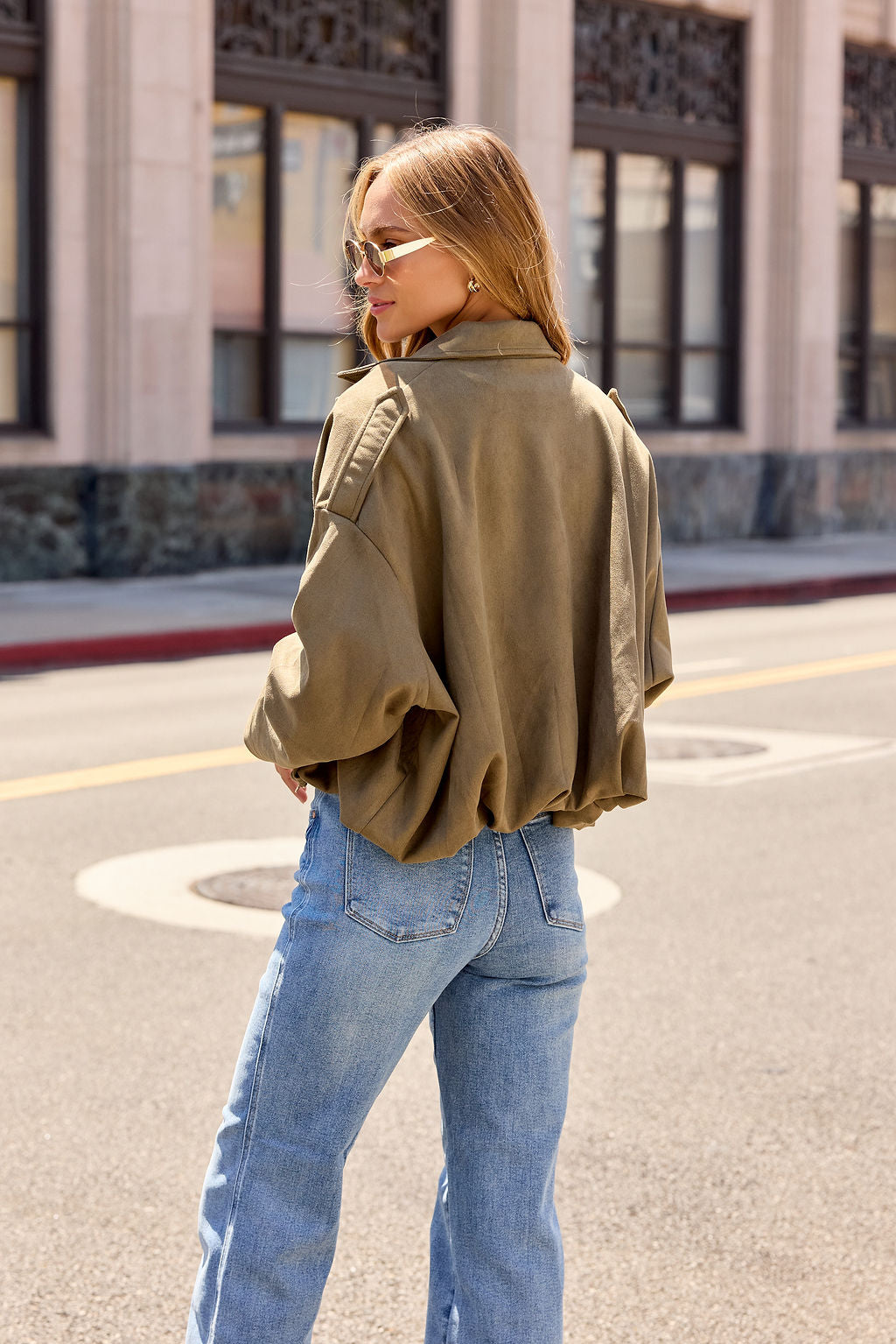 Woman in sunglasses and jeans wears the Suede Bubble Hem Jacket - Olive, standing on a sunny city street, looking over her shoulder.