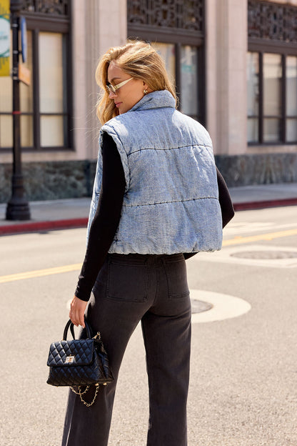 Woman with long hair wears fall layers: a black top, pants, and Vintage Havana Puffer Vest - Denim, holding a black quilted purse.
