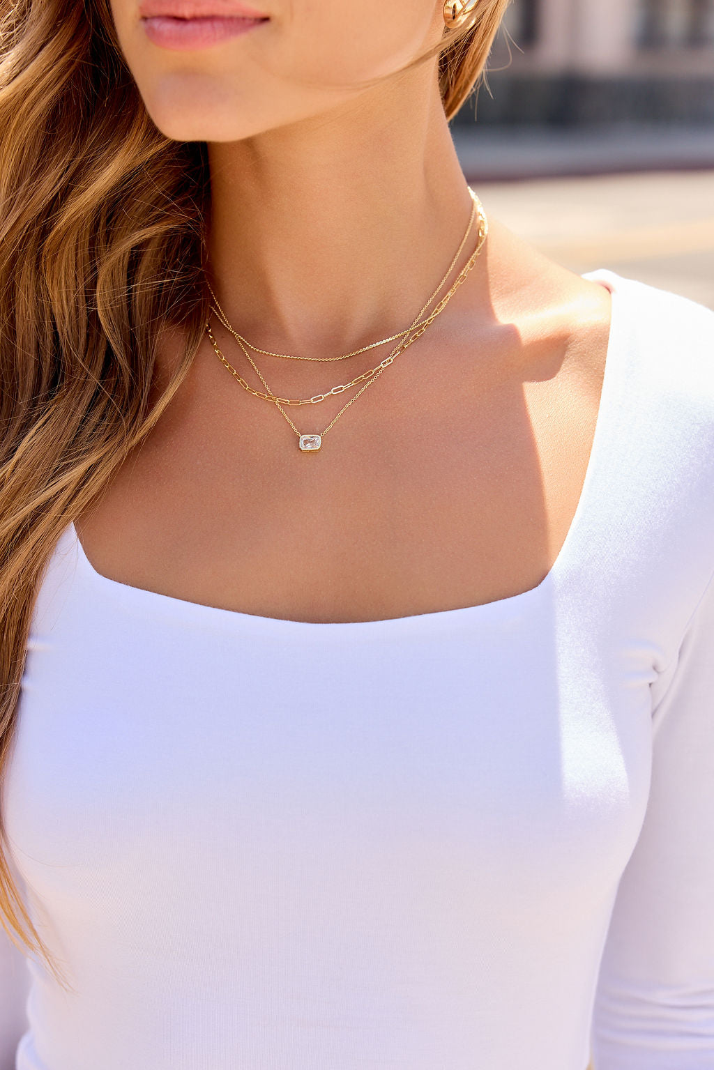 Woman in a white top wears the Layered Emerald Cut Necklace - Gold; long wavy hair, blurred outdoor background.