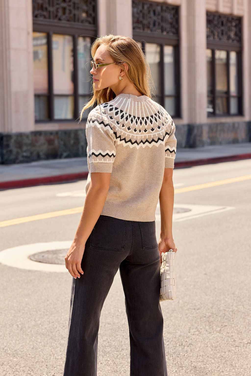 Woman in sunglasses wears a Taupe Aztec Print Sweater and dark pants, holding a white purse on a city street.