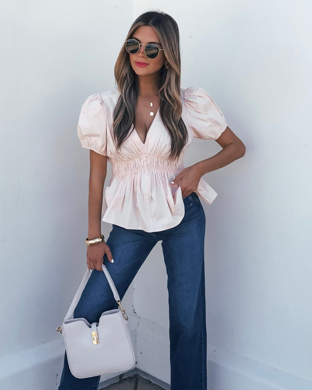 Woman wearing a light pink blouse with puff sleeves and blue jeans, holding a white handbag against a white background.