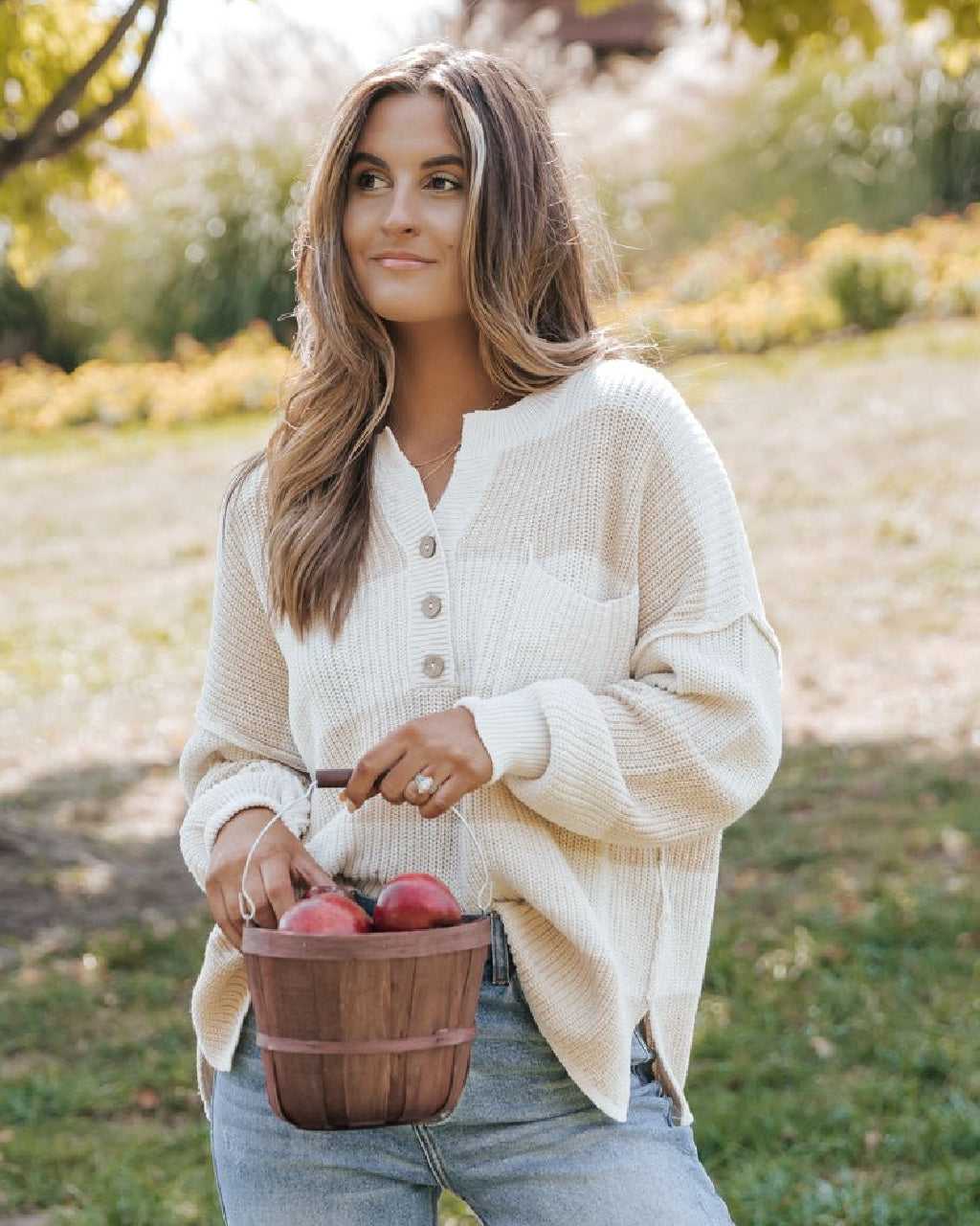 A woman wearing a cream-colored waffle-knit button-up cardigan and light blue jeans stands outdoors in a park holding a wooden basket filled with red apples.