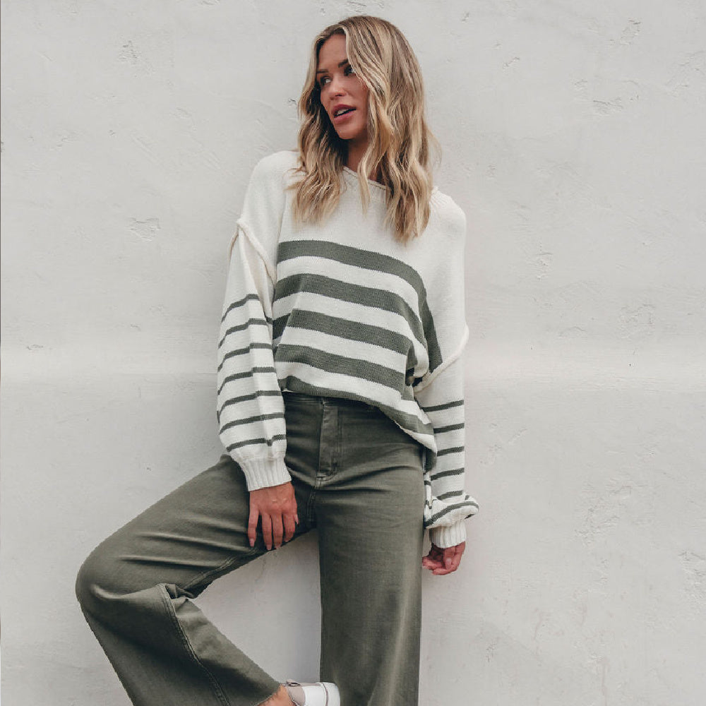 A woman wearing a cream and olive green striped knit sweater and olive green wide-leg pants, leaning against a textured white wall.