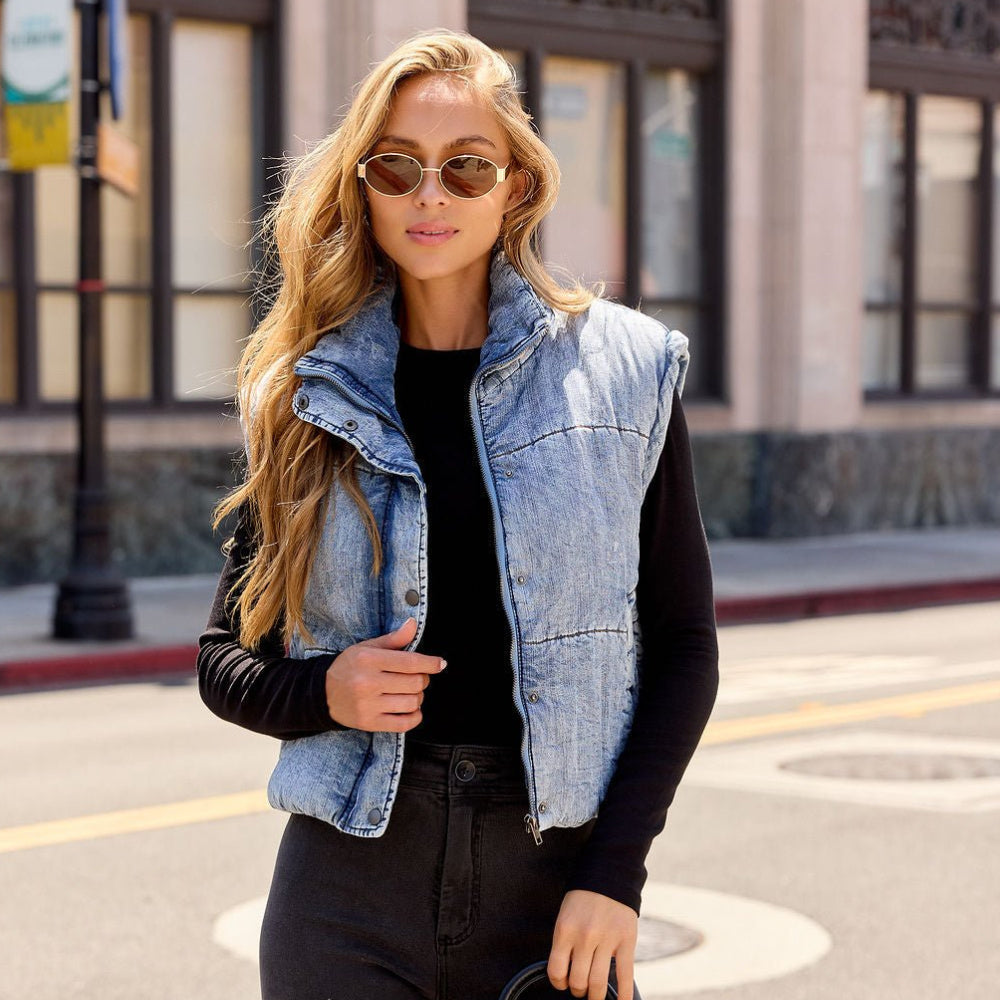 A woman wearing a light blue denim sleeveless puffer vest over a black long-sleeve top and black jeans, standing on a city street.