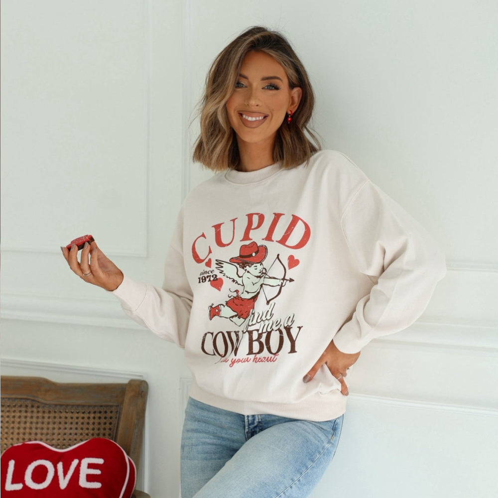Hayley A is standing indoors against a wall wearing our Light_Beige_Cupid_Cowboy_Graphic_Sweatshirt