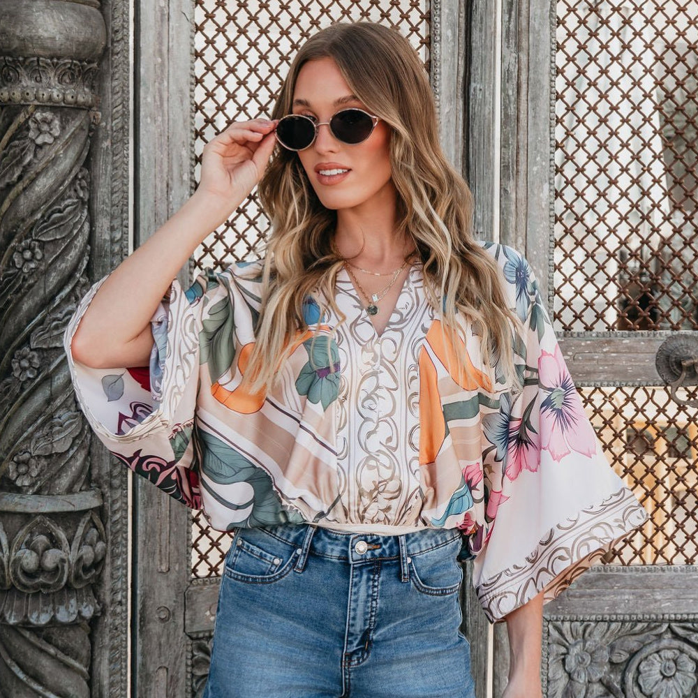 Woman wearing a colorful floral kimono over a patterned top and jeans, standing in front of a textured wall.