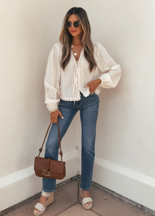 Woman wearing a white blouse, blue jeans, and carrying a brown handbag against a plain wall.