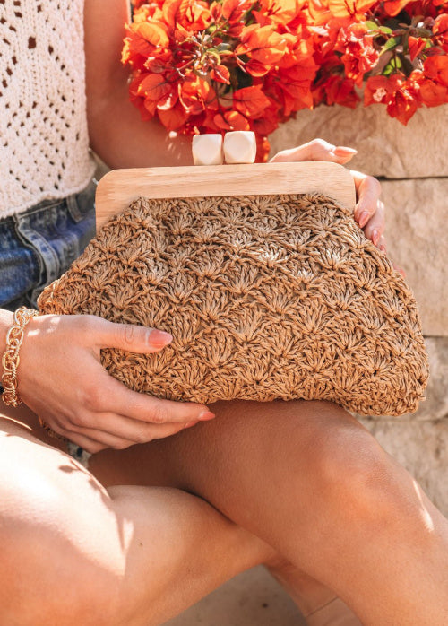 Person holding a woven clutch with a stone wall and flowers in the background