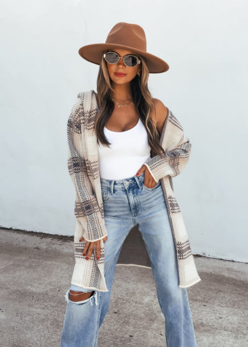 A woman wearing a brown wide-brim fedora hat, round sunglasses, a white tank top, light-wash distressed jeans, and an open beige-and-brown plaid cardigan, standing against a plain white wall.