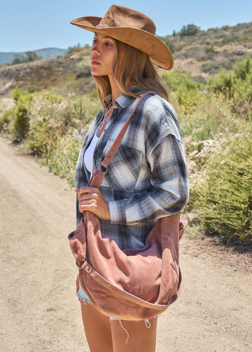 Woman in a plaid shirt and cowboy hat standing on a dirt road with a pink bag.