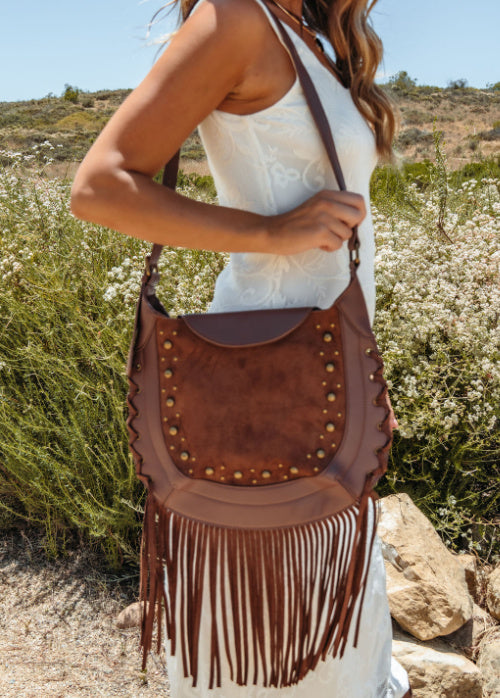 Person holding a brown leather handbag with fringe in a natural setting