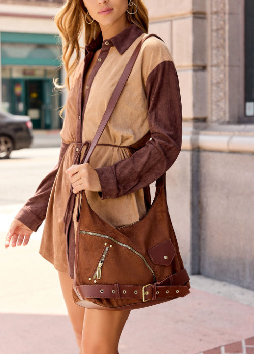 A woman wearing a color-block brown and tan long-sleeve shirt dress, carrying a matching brown suede-look shoulder bag with a strap and belt detail, standing outdoors on a city sidewalk.