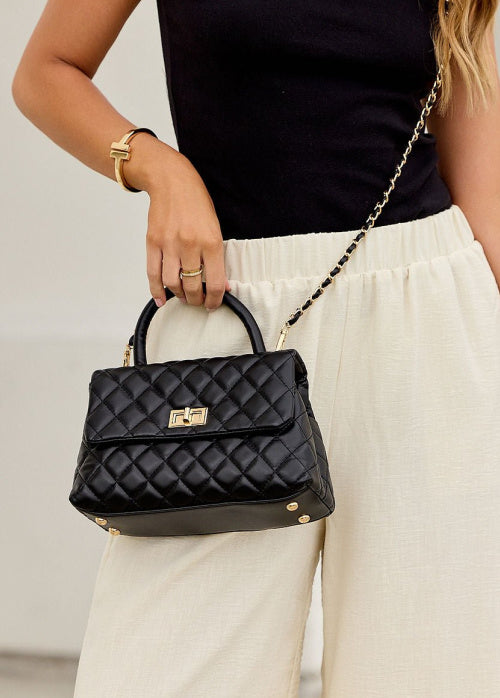 A woman wearing a black sleeveless top and cream wide-leg pants holds a black quilted top-handle handbag with gold-tone hardware and a chain strap.