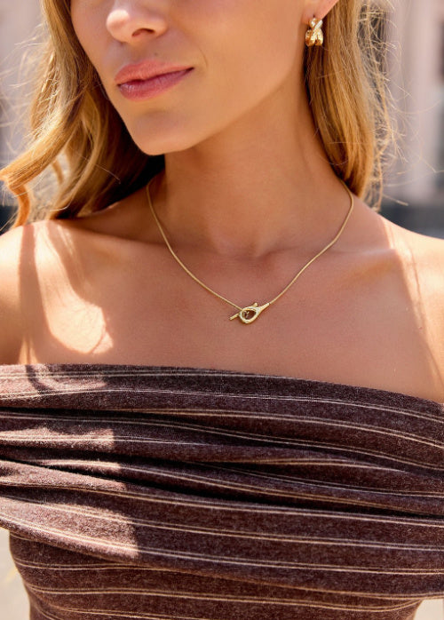 A close-up lifestyle shot of a woman wearing a delicate gold necklace with a small openwork heart pendant, paired with a brown ribbed strapless top.