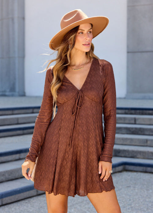 A woman wearing a brown long-sleeve mini dress with a tie-front detail, styled with a wide-brim tan hat, standing outdoors in front of gray stone steps.