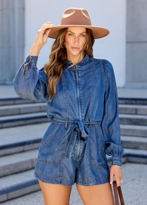 A woman wearing a blue denim romper with a front zipper, long sleeves, and a matching waist tie, styled with a tan wide-brim hat in front of outdoor stone steps.