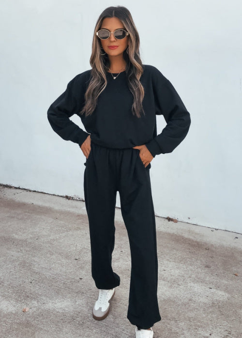 A woman wearing a black long-sleeve sweatshirt and matching black wide-leg sweatpants, styled with white sneakers and sunglasses, standing against a white wall.